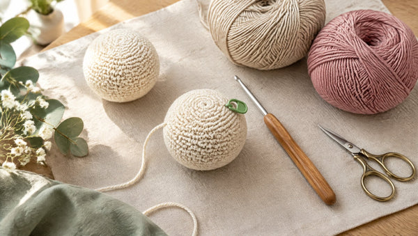 Two cream crocheted amigurumi balls on linen beside a wooden crochet hook, cotton yarn and a green stitch marker