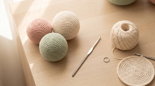 Three smooth round crocheted amigurumi balls in pastel yarn with a crochet hook and yarn on a cozy craft desk