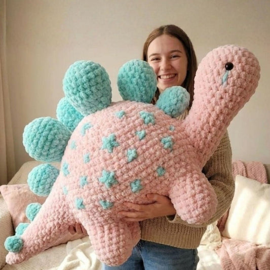 Giant pink and turquoise crochet stegosaurus dinosaur amigurumi held by smiling woman showing finished pattern result